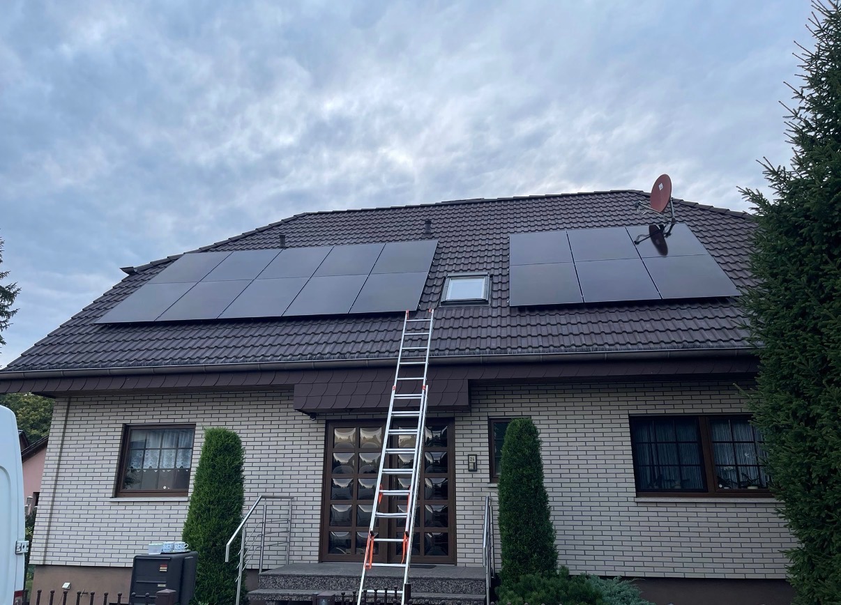 Panele słoneczne na brązowym dachu domu w Glinno, widoczne z ziemi. Aluminiowa drabina oparta o dach. Antena satelitarna. Pochmurne niebo.