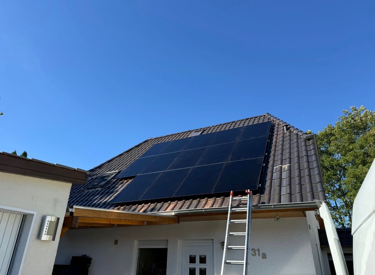 Panele fotowoltaiczne na dachu domu jednorodzinnego, drabina oparta o dach, błękitne niebo w tle. Widok z dołu, częściowo widoczny biały samochód dostawczy.