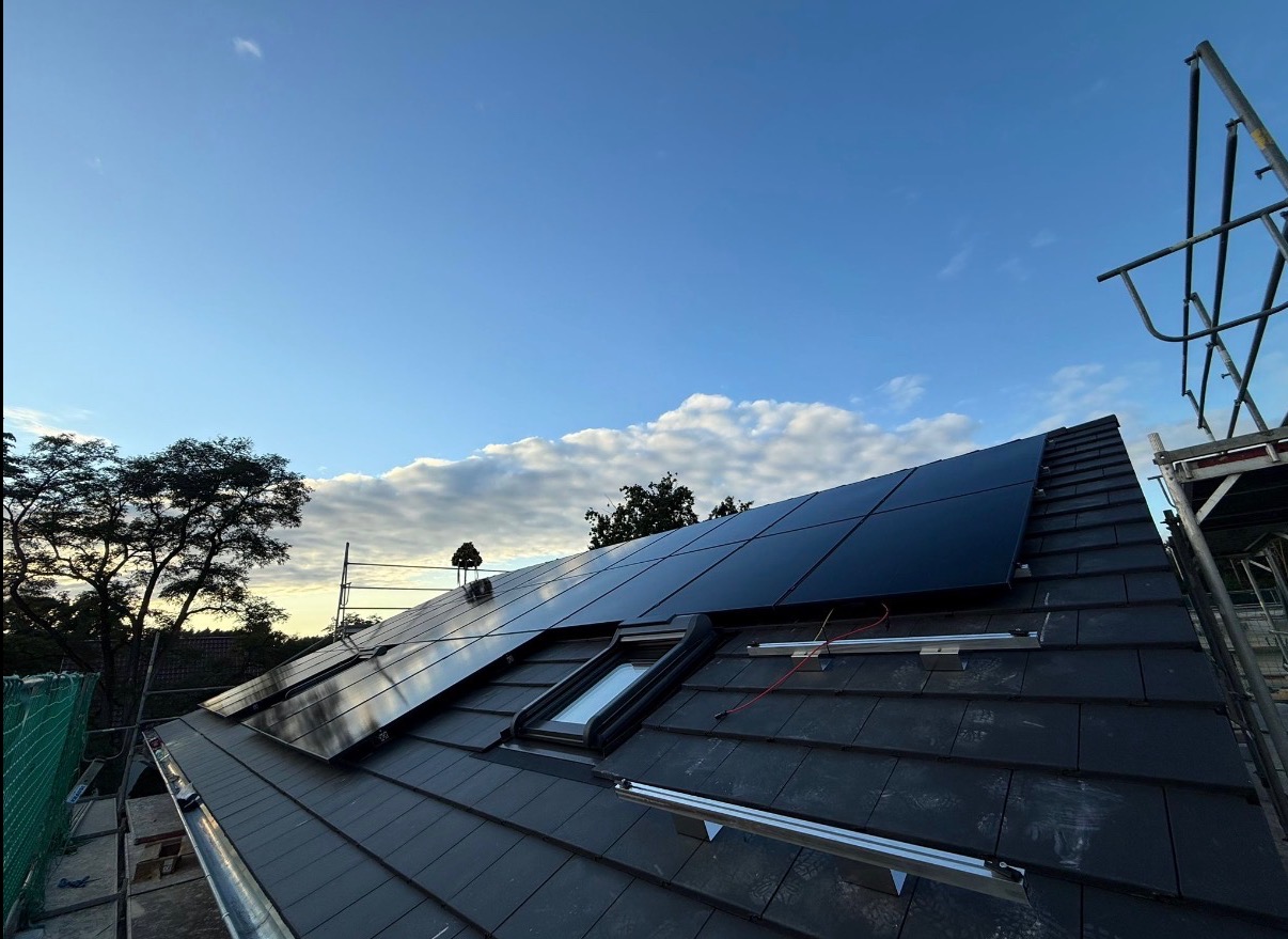 Czarne panele słoneczne na dachu z dachówką, widok z dołu, częściowo widoczne rusztowanie, błękitne niebo z chmurami w tle, wczesny etap montażu.