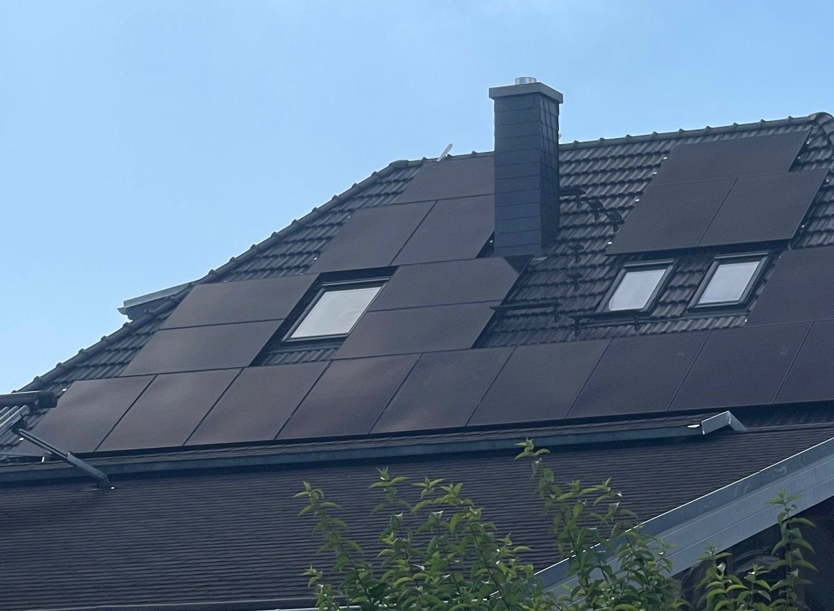 Ciemne panele słoneczne na dachu domu z oknami dachowymi i kominem, widok z dołu, częściowo zasłonięte przez zielone krzewy.
