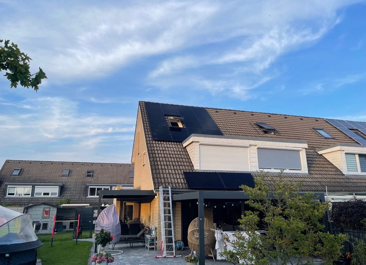 Instalacja paneli na dachu domu jednorodzinnego z widoczną drabiną i częściowo zamontowanymi panelami. Widok na dach i fragment ogrodu.