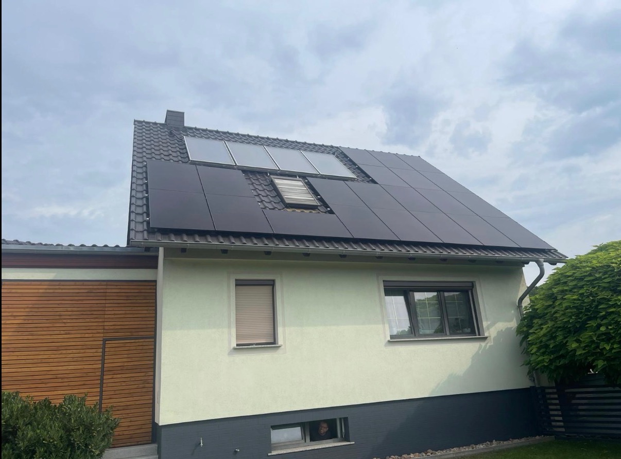 Dom z panelami słonecznymi na dachu, widok z dołu. Część paneli jest w kolorze czarnym, a część w kolorze szarym. Widoczne okno dachowe i fragment ogrodu.