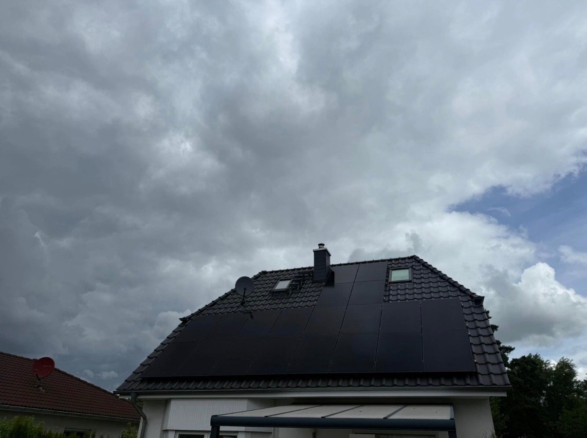 Czarne panele słoneczne na dachu domu z kominem i oknami dachowymi, na tle pochmurnego nieba. Widoczna antena satelitarna i fragment sąsiedniego budynku.