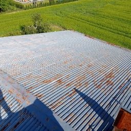 KAMBUD Kamil Warchał - Blaszany dach z widocznymi śladami rdzy i cieniem osoby robiącej zdjęcie na tle zielonego pola, sfotografowany z góry w słoneczny dzień.