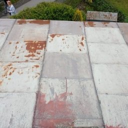 KAMBUD Kamil Warchał - Blaszany dach z widocznymi śladami rdzy i ubytkami farby, sfotografowany z góry pod kątem. W tle zieleń drzew i krzewów, oraz fragmenty postaci ludzkich.