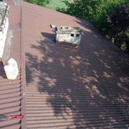 KAMBUD Kamil Warchał - Ciemnobrązowy dach z blachy falistej, widoczny komin z cegły, narzędzia i pojemnik z płynem. Cień drzew na powierzchni dachu.
