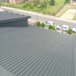 Szary, blaszany dach po renowacji, widok z góry na tle zabudowań i ulicy z zaparkowanymi autami. Charakterystyczne, podłużne przetłoczenia blachy.