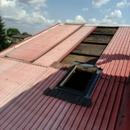 Czerwony dach z blachy trapezowej w trakcie montażu włazu dachowego. Widoczne deski pod blachą i fragment nieba z chmurami. Ujęcie z góry.