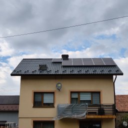 KAMBUD Kamil Warchał - Dwupiętrowy dom z nowym, ciemnym dachem z dachówki, na którym zamontowano czarne panele fotowoltaiczne i okno dachowe. Beżowa elewacja, balkon z czarną balustradą częściowo osłonięty folią,...