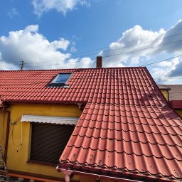 KAMBUD Kamil Warchał - Czerwony dach z blachodachówki na żółtym budynku mieszkalnym, z zamontowanym oknem dachowym i białymi rynnami. Widoczne drobne ślady zużycia pokrycia.