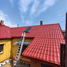 KAMBUD Kamil Warchał - Świeżo odnowiony czerwony dach z falistej blachy na żółtym budynku, z drabiną opartą o ścianę i folią ochronną na oknie, pod błękitnym niebem.