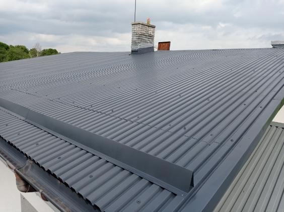 Szary, blaszany dach z widocznymi rynnami i kominem z cegły w tle, sfotografowany pod kątem. Widoczne delikatne obłoki na niebie.