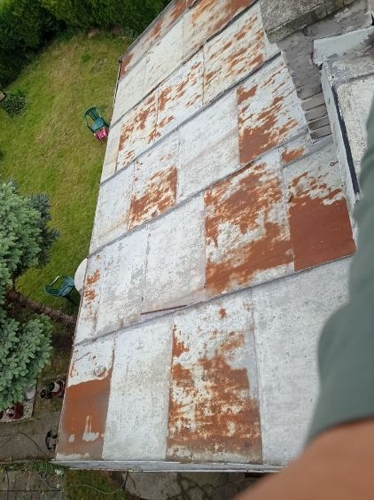 Zardzewiały dach z blachy falistej, widok z góry na fragment dachu z wyraźnymi śladami korozji i łuszczącą się farbą, na tle zielonego trawnika i drzew.