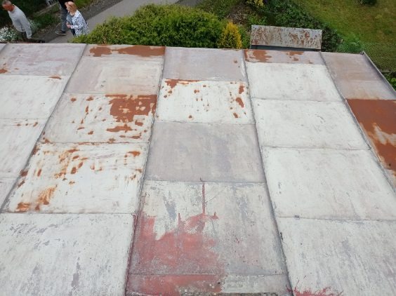 Blaszany dach z widocznymi śladami rdzy i ubytkami farby, sfotografowany z góry pod kątem. W tle zieleń drzew i krzewów, oraz fragmenty postaci ludzkich.