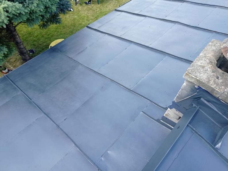 Metalowy dach po renowacji, widok z góry, z fragmentem komina i zielonym drzewem w tle. Blacha w odcieniach szarości, równo ułożona. Świeżo pomalowana powierzchnia.