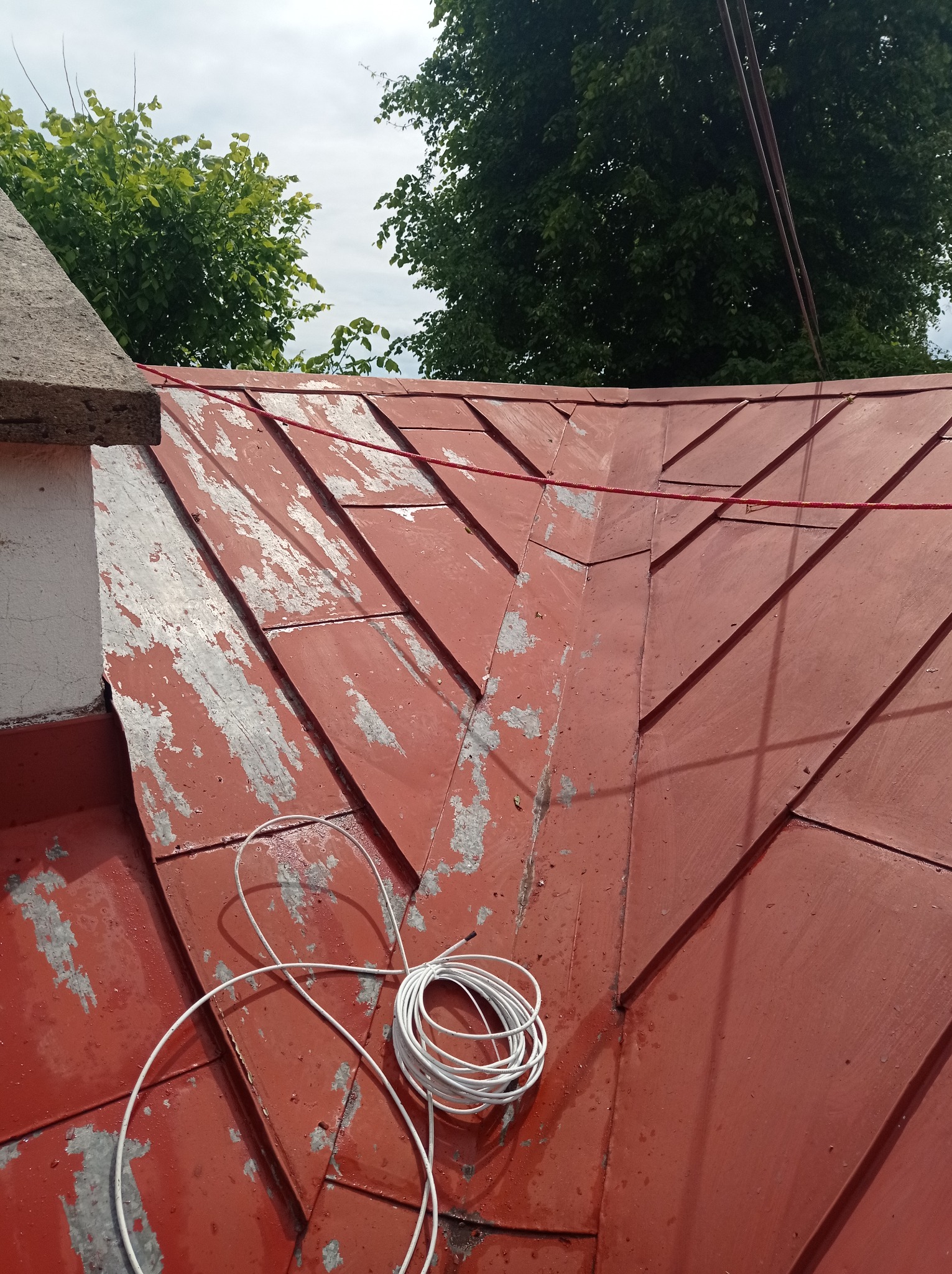 Czerwony dach z blachy trapezowej, miejscami łuszcząca się farba, zwój białego kabla na powierzchni, komin i drzewa w tle.