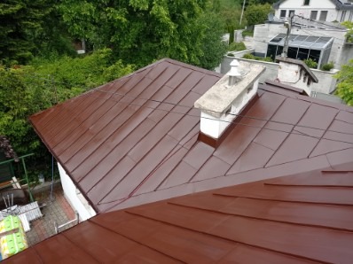 Ciemnobrązowy, metalowy dach po renowacji, widok z góry. W tle zieleń drzew i zabudowa mieszkalna. Widoczny komin z betonową obudową.