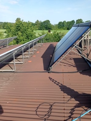 Brązowy dach z panelami słonecznymi w rzędach, z pojedynczym panelem po prawej. W tle zieleń drzew i błękit nieba. Widoczne kable na dachu.