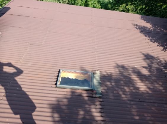 Ciemnobrązowy dach z blachy falistej z zamontowanym oknem dachowym; cień osoby robiącej zdjęcie widoczny w lewym dolnym rogu.