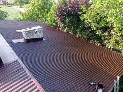 Ciemnobrązowy, blaszany dach z ceglanym kominem, otoczony zielenią drzew. Widoczne narzędzia malarskie przy krawędzi dachu. Ujęcie z góry, w słoneczny dzień.