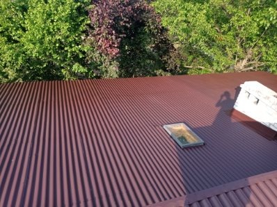 Ciemnobrązowy dach z blachy trapezowej po renowacji, widok z góry. Na dachu lukarna i cień osoby. W tle zieleń drzew.