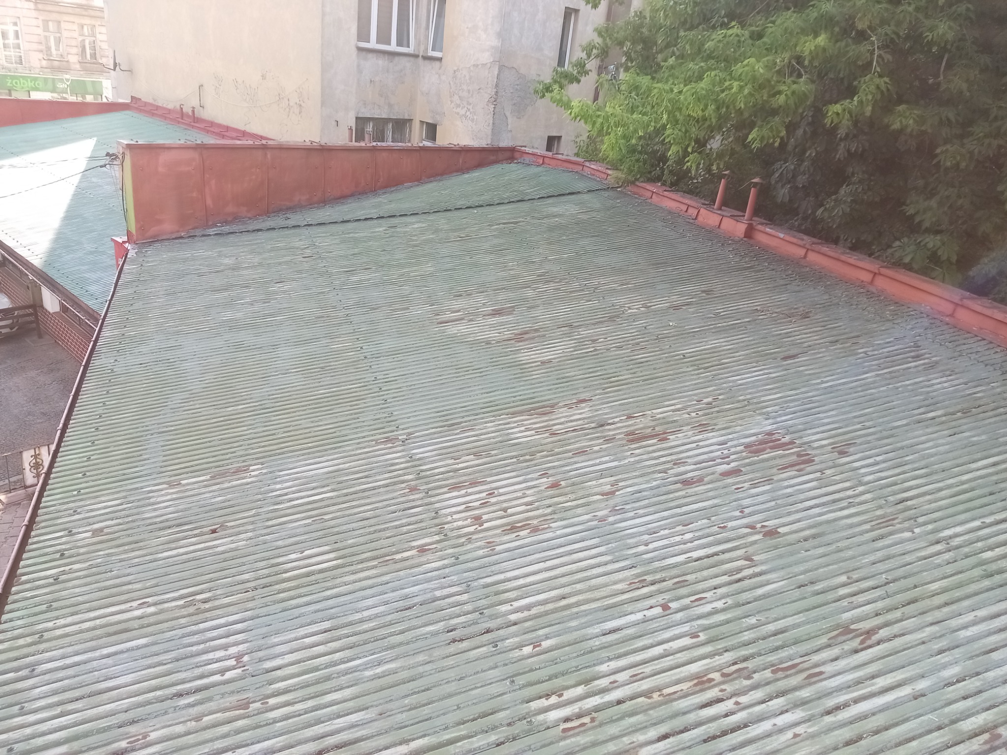 Falista blacha na dachu budynku z widocznymi śladami korozji i miejscami odchodzącej farby, z fragmentami rynien w kolorze czerwonym. Widok z góry.