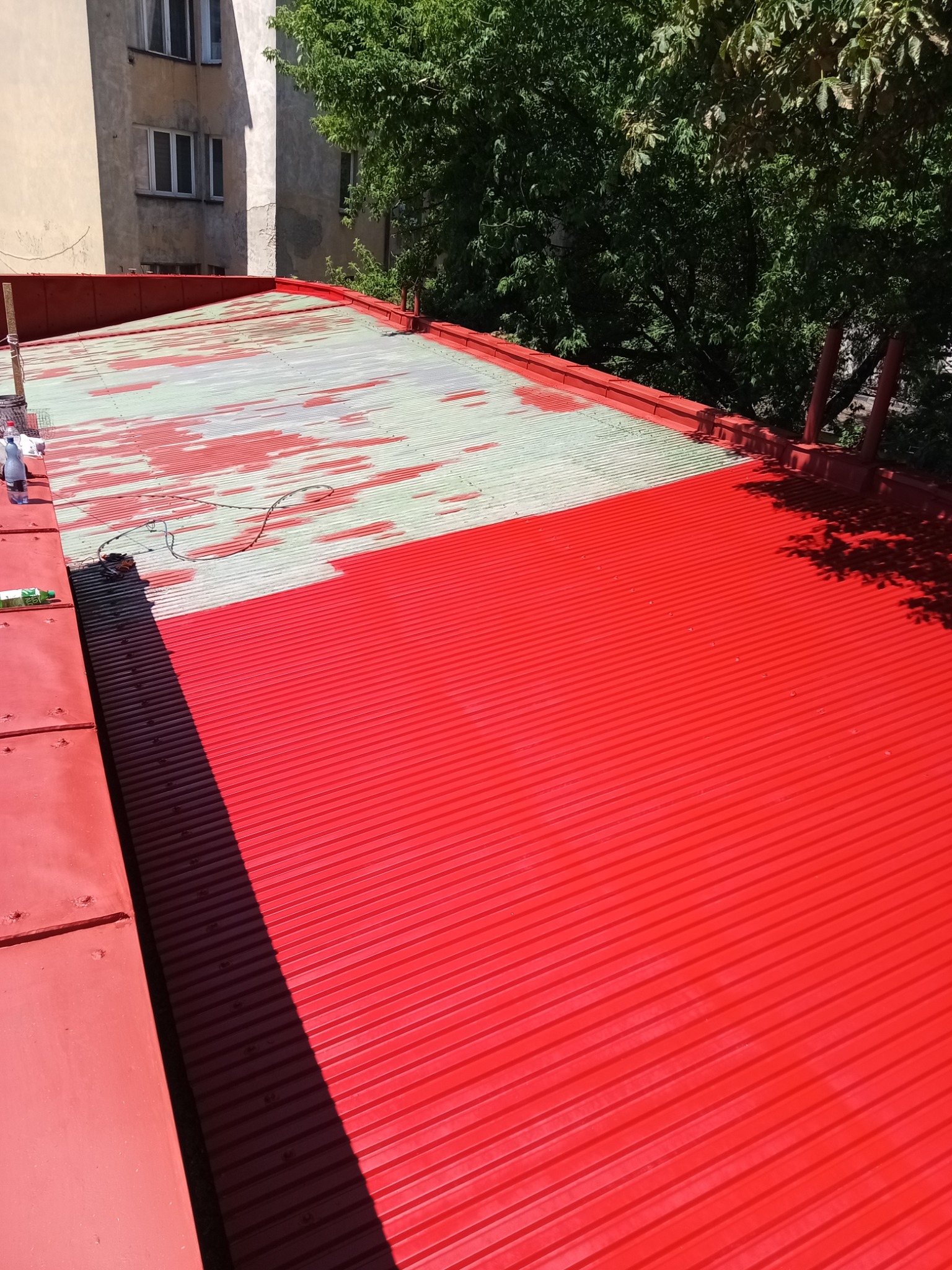 Częściowo pomalowany na czerwono blaszany dach budynku. Widoczne ślady wcześniejszej powłoki i butelka wody. Tło: budynek i drzewa.