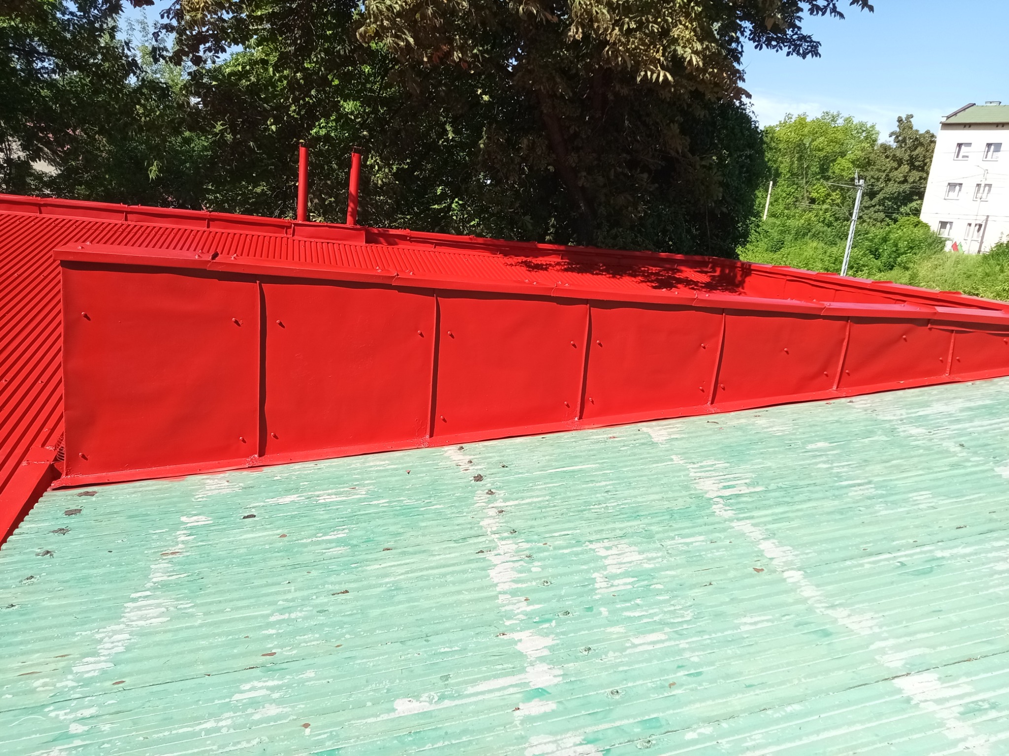 Czerwony dach z blachy falistej po malowaniu, widoczne detale łączeń i faktura powierzchni. Tło z zieleni drzew i fragment budynku.