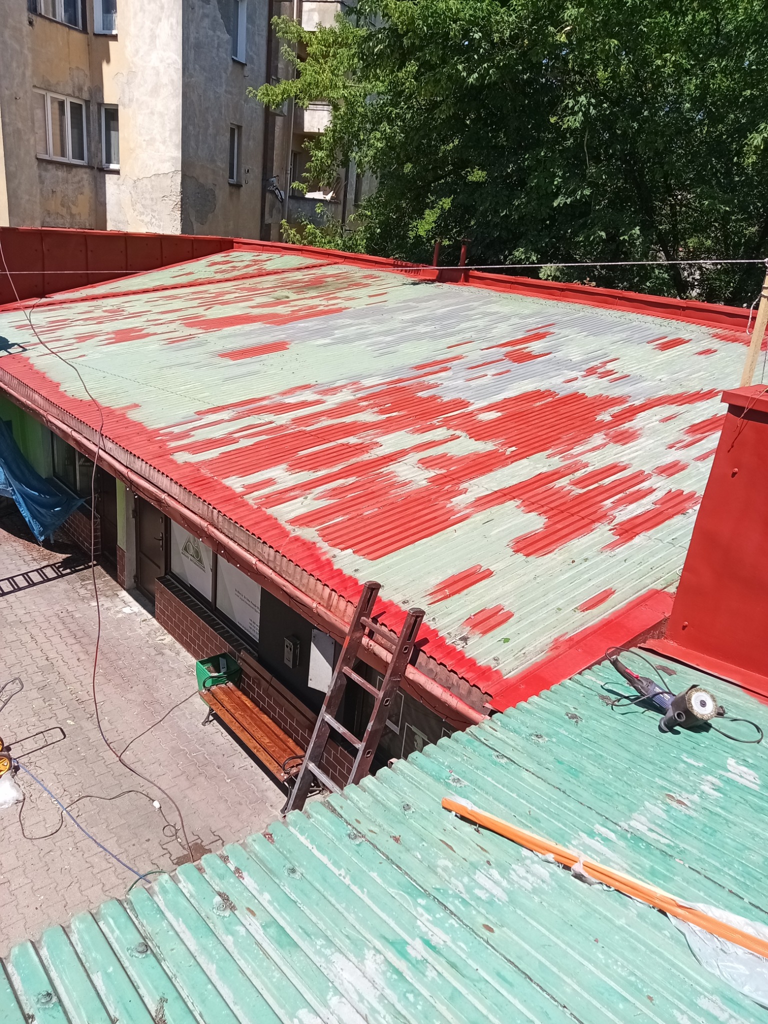 Częściowo pomalowany, blaszany dach z widocznymi śladami rdzy i starej farby. Na dachu leżą narzędzia i drabina. Widok z góry na budynek i drzewa w tle.