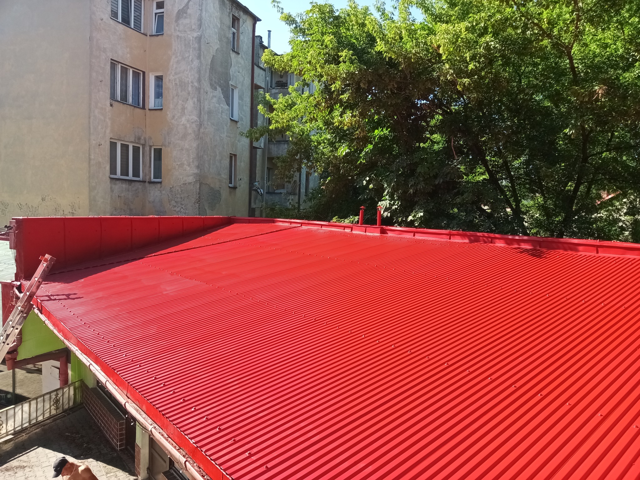 Czerwony dach z blachy falistej po malowaniu, widok z góry na tle budynku i zieleni. Świeża, intensywna barwa dachu wyróżnia się na tle otoczenia.