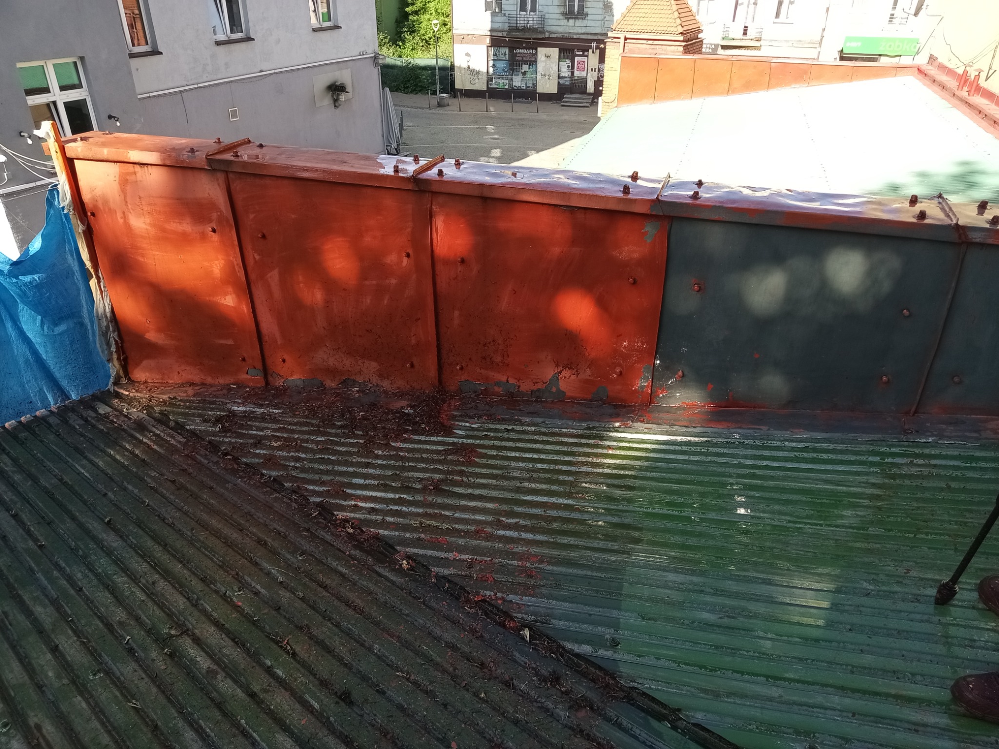 Częściowo pomalowany, pofałdowany dach z blachy trapezowej, z widoczną starą farbą i fragmentem niebieskiej folii ochronnej. W tle budynki miejskie.