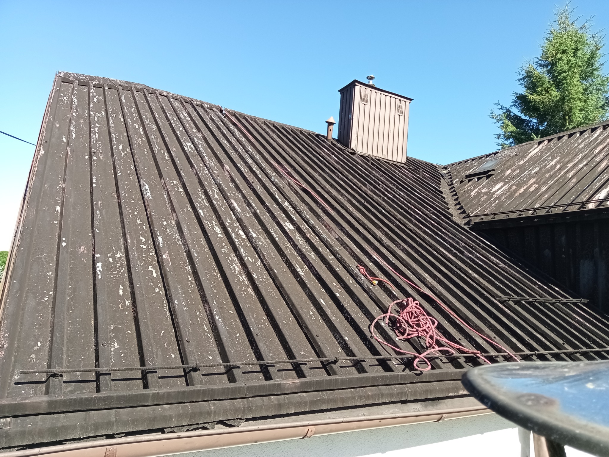 Pofalowany, metalowy dach z kominem i fragmentem rynny, pokryty łuszczącą się farbą. Widoczna lina zabezpieczająca. Ujęcie z dołu, w tle błękitne niebo i drzewo.