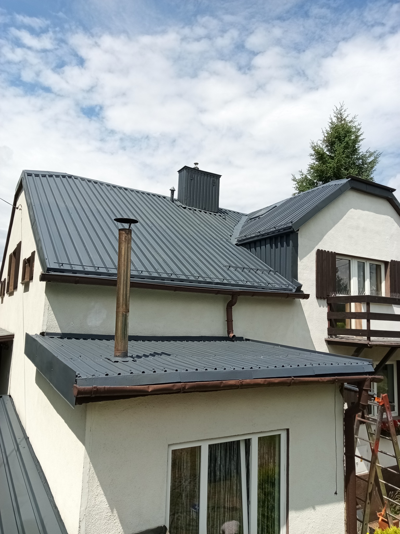 Szary, blaszany dach po renowacji na tle błękitnego nieba z chmurami. Widoczne rynny, komin i fragment elewacji domu. Ujęcie z dołu.