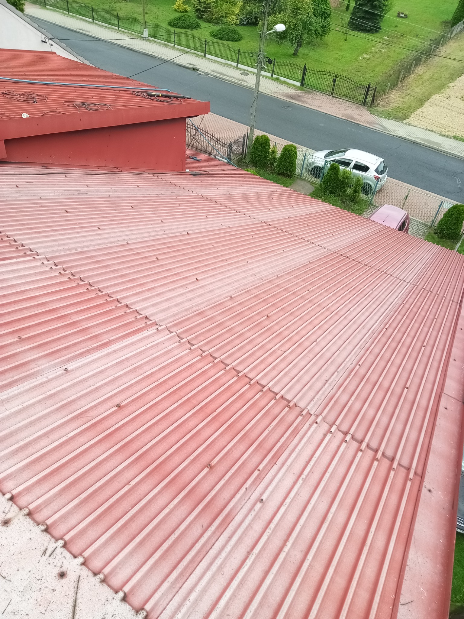 Czerwony dach z blachy trapezowej, widoczny z góry, z elementami instalacji na krawędzi. W tle ulica i zieleń.