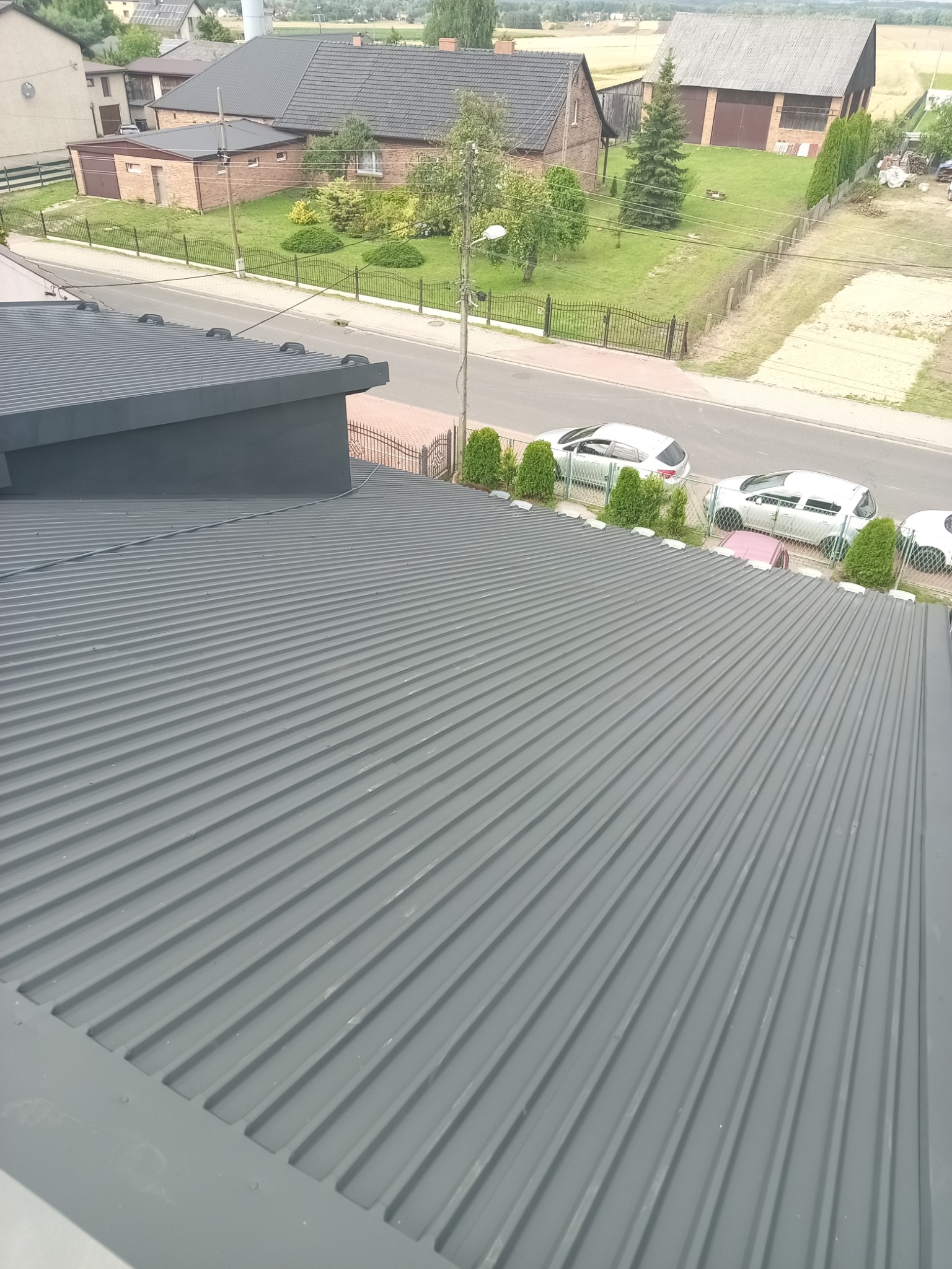 Szary, blaszany dach po renowacji, widok z góry na tle zabudowań i ulicy z zaparkowanymi autami. Charakterystyczne, podłużne przetłoczenia blachy.