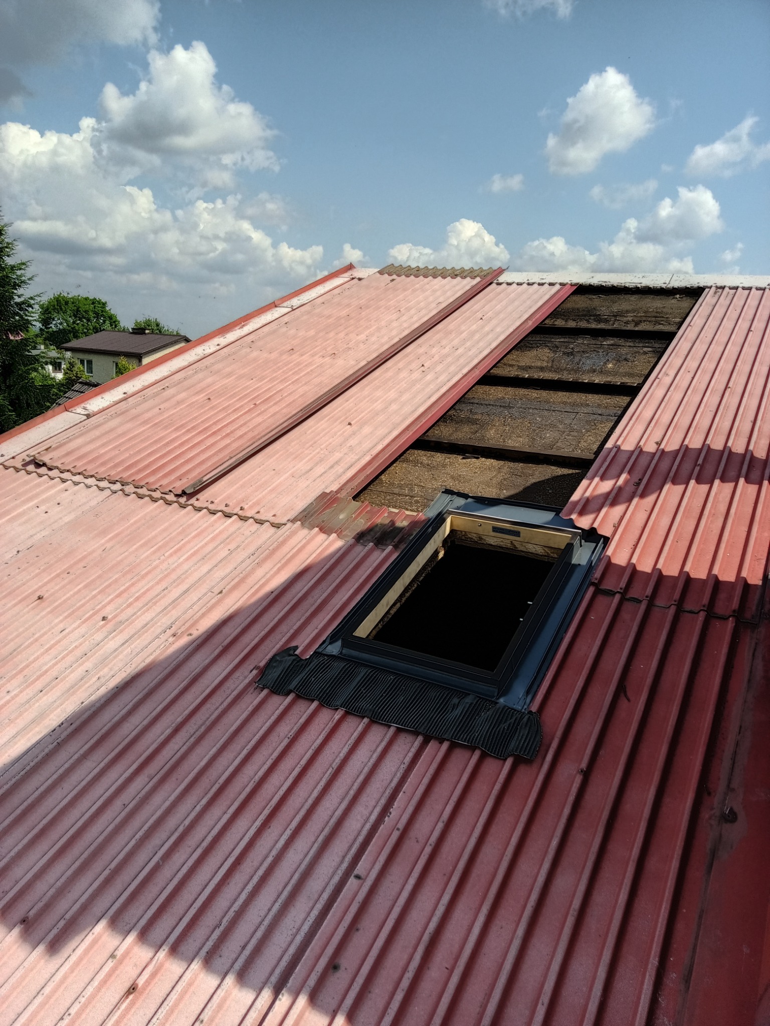 Czerwony dach z blachy trapezowej w trakcie montażu włazu dachowego. Widoczne deski pod blachą i fragment nieba z chmurami. Ujęcie z góry.
