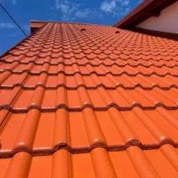 Czerwona dachówka ceramiczna, świeżo po renowacji, błyszcząca w słońcu pod błękitnym niebem. Widok z dołu ukazuje regularny wzór dachówek i fragment orynnowania.