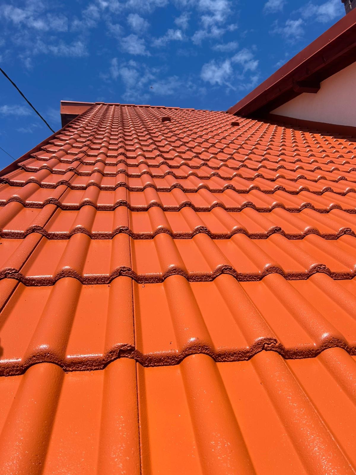 Czerwona dachówka ceramiczna, świeżo po renowacji, błyszcząca w słońcu pod błękitnym niebem. Widok z dołu ukazuje regularny wzór dachówek i fragment orynnowania.