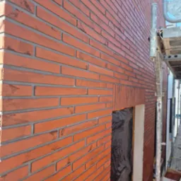 Elewacja budynku wykończona czerwoną cegłą klinkierową, widoczne fragmenty rusztowania i zabezpieczone folią okno.