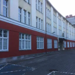 Budynek z czerwoną elewacją na parterze i kremową powyżej, białe ramy okienne z kratami, kostka brukowa na chodniku, widoczne oznakowanie parkingu dla niepełnosprawnych.