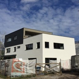 PROFREM DARIUSZ DOMACHOWSKI - Nowoczesny dwukolorowy dom z płaskim dachem na etapie wykończenia elewacji, widoczny zza tymczasowego ogrodzenia i samochodu dostawczego z logo firmy budowlanej.