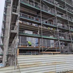 Wielopiętrowy budynek w trakcie prac związanych z izolacją termiczną elewacji, widoczne rusztowania, płyty izolacyjne z charakterystycznymi białymi kołkami mocującymi, pracownik w żółtej kamizelce...