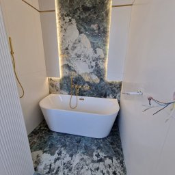 PROFREM DARIUSZ DOMACHOWSKI - Nowoczesna łazienka z białą wanną wolnostojącą, złotą armaturą i marmurową ścianą z podświetleniem LED, widoczne niedokończone prace elektryczne.