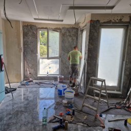 PROFREM DARIUSZ DOMACHOWSKI - Wnętrze w trakcie prac wykończeniowych, widoczne marmurowe płyty wokół okien, rozłożone narzędzia i drabina, podłoga wyłożona błyszczącymi płytkami imitującymi marmur.