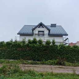 PROFREM DARIUSZ DOMACHOWSKI - Dom jednorodzinny z szarą blachodachówką, częściowo zasłonięty wysokim żywopłotem, z widocznymi rusztowaniami przy ścianie szczytowej, wskazującymi na prace elewacyjne.