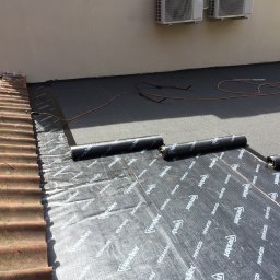 PROFREM DARIUSZ DOMACHOWSKI - Montaż hydroizolacji na dachu budynku z widocznymi rolkami papy bitumicznej Nexler i palnikiem dekarskim, fragment dachu pokryty starą dachówką