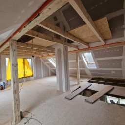 PROFREM DARIUSZ DOMACHOWSKI - Adaptacja poddasza: surowe wnętrze z drewnianą konstrukcją stropu, widoczne płyty OSB, okna dachowe, ściany przygotowane do wykończenia, narzędzia i materiały budowlane w tle.