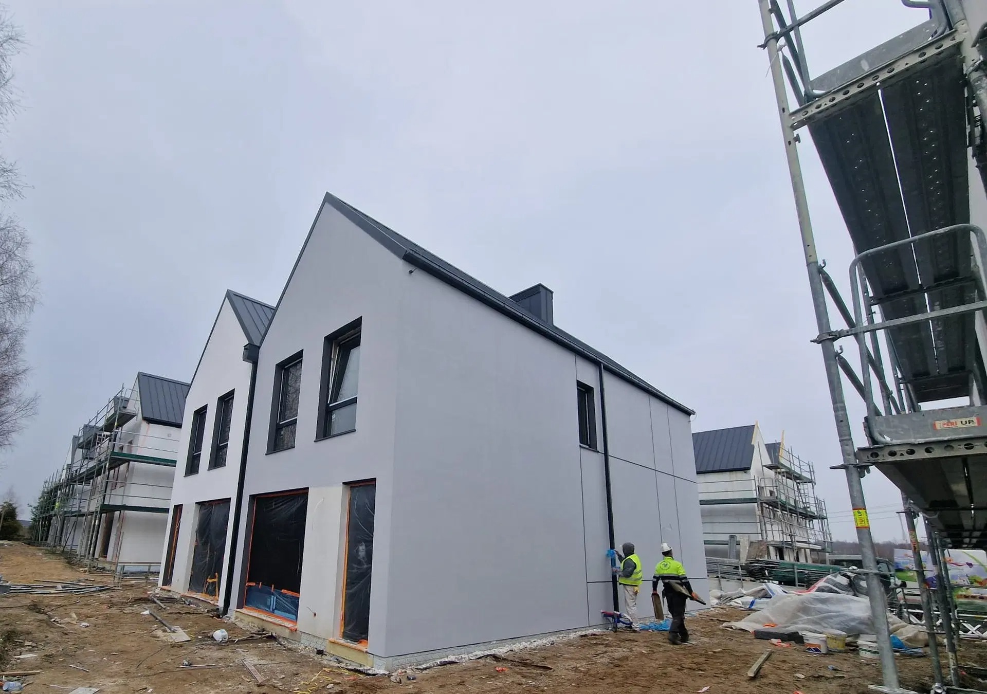 Docieplanie i malowanie elewacji osiedla domków jednorodzinnych w stylu nowoczesnym; widoczne rusztowania, pracownicy w odblaskowych kamizelkach i świeżo otynkowana szara elewacja.