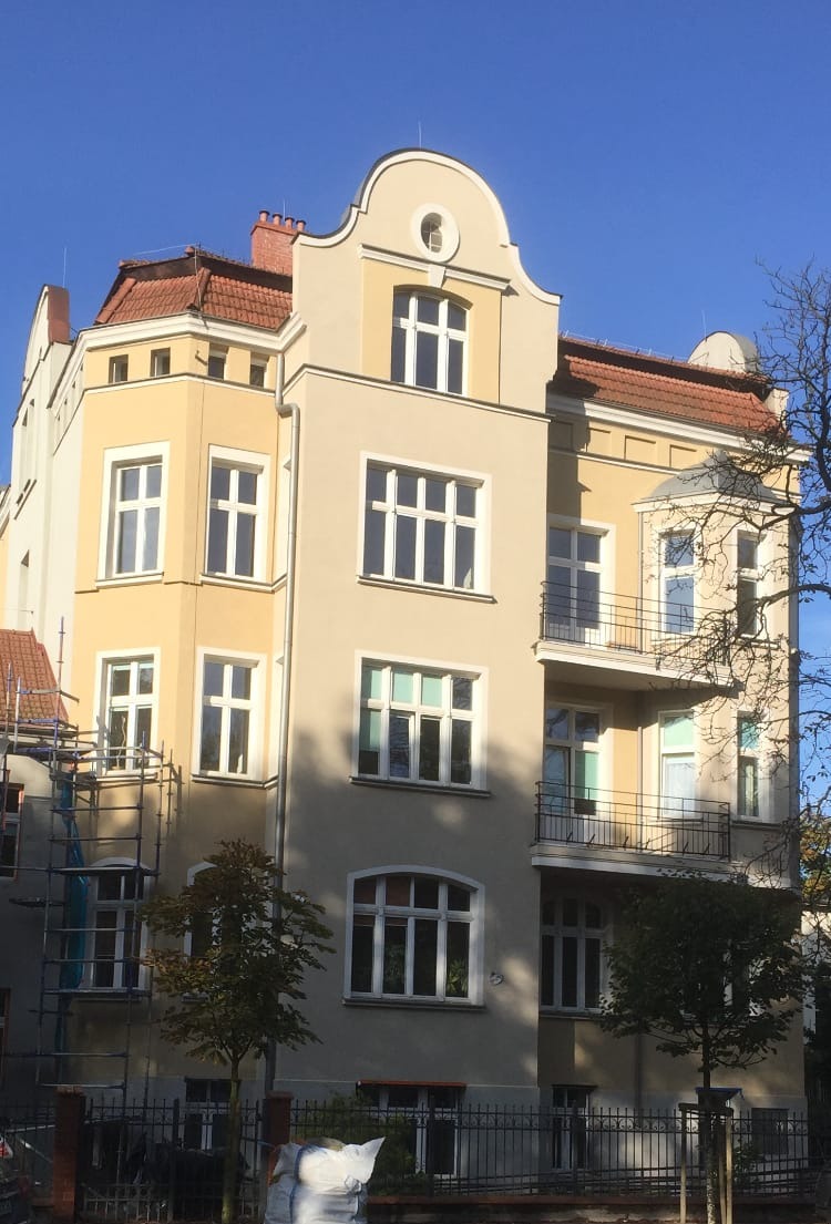 Odnowiona kamienica z jasnożółtą i beżową elewacją, widoczne rusztowania z boku budynku, błękitne niebo w tle.