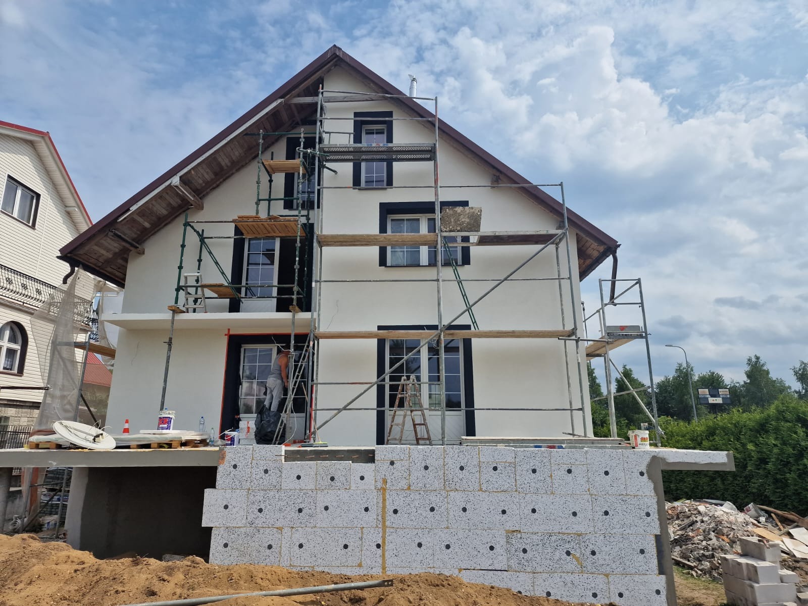 Dom jednorodzinny w trakcie prac elewacyjnych, widoczne rusztowania, ocieplenie fundamentów styropianem oraz pracownik na drabinie przy oknie.